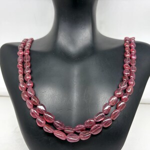Puede incluir: Un collar de tres hilos con cuentas rosas ovaladas y translúcidas. Las cuentas están ensartadas, creando un efecto de capas. El collar se exhibe en un maniquí negro, mostrando el diseño y el color de la joya.