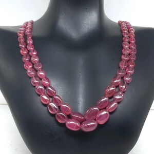 May include: A double-strand necklace featuring oval-shaped, pinkish-red gemstones. The necklace is displayed on a black mannequin bust, showcasing the polished stones' color and shape. The beads are strung together, creating a layered effect.