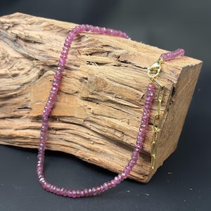 May include: A necklace composed of small, faceted, deep pink gemstone beads. The strand features a gold-toned metal lobster claw clasp and a short length of gold-toned oval link chain for adjustment, creating a delicate and refined jewelry piece.