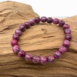 May include: A stretch bracelet composed of round, polished beads in shades of magenta, deep pink, and purple, featuring natural dark speckles and inclusions throughout each stone. The beads are strung on a flexible cord to form a circular shape.