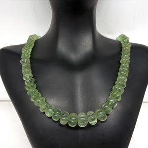 May include: A light green beaded necklace displayed on a black mannequin. The necklace features rounded, translucent beads with a ribbed design. The beads are strung together to form a necklace. The necklace is a light green color.