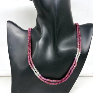 Puede incluir: Un collar de varias capas con pequeñas gemas rosas facetadas y cuentas transparentes. El collar se exhibe en un busto de maniquí negro. Las gemas tienen un color profundo y rico, y las cuentas transparentes añaden un toque de brillo.