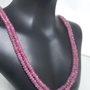 May include: A triple-strand necklace composed of small, faceted pink gemstones. The necklace is draped on a black mannequin, showcasing the color and texture of the beads. The gemstones have a subtle sparkle, reflecting light.
