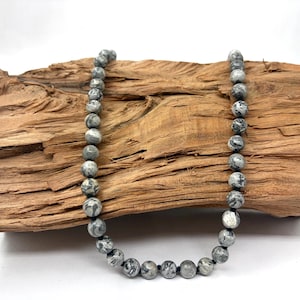 Può includere: Una collana composta da perline sferiche caratterizzate da un motivo marmorizzato nei toni del grigio, bianco e nero. Le perline sono infilate su un cordino scuro, creando una disposizione circolare continua con una superficie liscia e lucida.