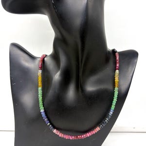 Peut inclure: Un collier de pierres précieuses multicolores présenté sur un mannequin noir. Le collier présente un dégradé de couleurs, notamment rouge, jaune, vert, bleu et noir. Les pierres précieuses sont petites et facettées, créant un effet scintillant.