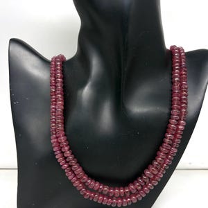 May include: A three-strand necklace composed of small, round, reddish-pink gemstones. The necklace is displayed on a black mannequin bust against a white background. The gemstones appear to be uniform in size and shape.
