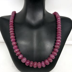 May include: A necklace of carved, deep pink gemstones displayed on a black mannequin. The beads are round with a ribbed texture, strung together to form a necklace. The colour is a rich, saturated pink.