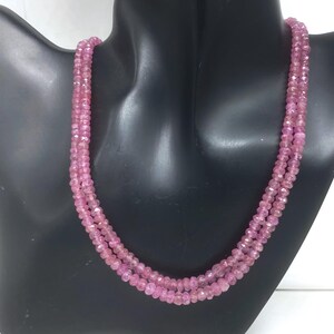 Puede incluir: Un collar de tres hilos compuesto por pequeñas piedras preciosas rosas facetadas. El collar se muestra sobre un busto de maniquí negro, destacando el diseño y el color. Las piedras preciosas son de tamaño y forma uniformes.