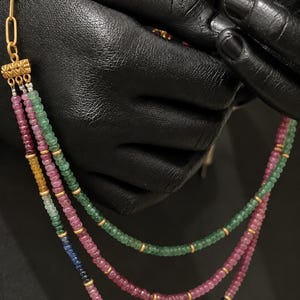 Puede incluir: Un collar de múltiples hilos con pequeñas cuentas de piedras preciosas facetadas en tonos rosa, verde, amarillo y azul. Los hilos están conectados por un espaciador de metal dorado con un patrón texturizado, unido a una cadena de eslabones.