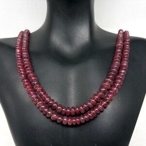 May include: A three-strand necklace composed of small, round, deep red beads. The beads are strung together, creating a layered effect. The necklace is displayed on a black mannequin bust, showcasing the jewelry's design and color.