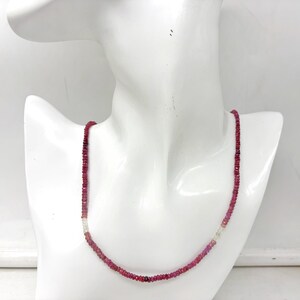 May include: A necklace composed of small, faceted, deep red gemstones. The necklace is displayed on a white mannequin bust. A few clear and pink stones are interspersed throughout the necklace.
