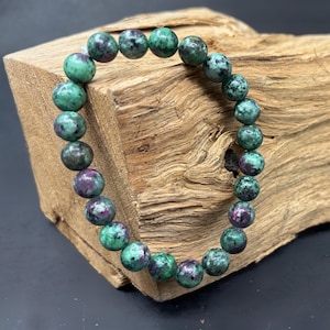 May include: A bracelet made of green and purple gemstone beads. The beads are round and strung together. The bracelet is displayed on a piece of wood. The gemstones are in shades of green and purple.