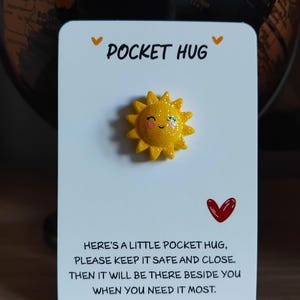 May include: A white card with the words "POCKET HUG" and a yellow sun with glitter. The card also has a red heart and text that reads "HERE'S A LITTLE POCKET HUG, PLEASE KEEP IT SAFE AND CLOSE. THEN IT WILL BE THERE BESIDE YOU WHEN YOU NEED IT MOST."