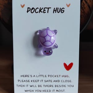May include: A lilac turtle charm on a white card with the words "POCKET HUG". The card has a message about keeping the charm safe and close. Decorative hearts are also present on the card.