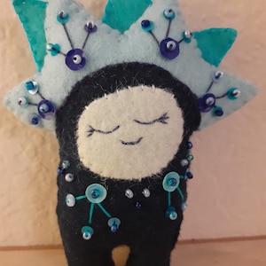 May include: A handmade felt doll with a serene expression. The doll features a light blue crown adorned with beads and sequins. The body is dark blue, embellished with beads and sequins. The face is white with embroidered eyes and a smile.