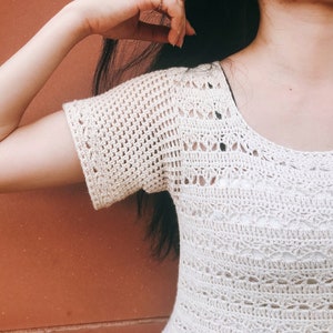 Puede incluir: Un top blanco de crochet con cuello redondo y mangas cortas. El top tiene un dise&ntilde;o delicado y calado.