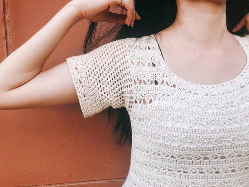 Puede incluir: Un top blanco de crochet con cuello redondo y mangas cortas. El top tiene un dise&ntilde;o delicado y calado.