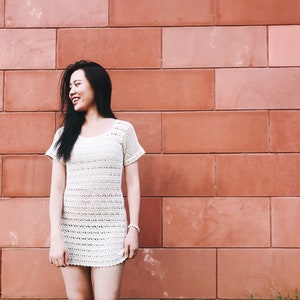 Puede incluir: Una mujer con el pelo largo y negro sonr&iacute;e y mira hacia la derecha. Lleva un vestido blanco de crochet y est&aacute; de pie frente a una pared de ladrillo rojo.