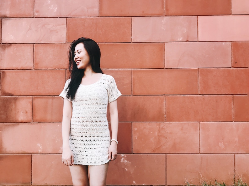Puede incluir: Una mujer con el pelo largo y negro sonr&iacute;e y mira hacia la derecha. Lleva un vestido blanco de crochet y est&aacute; de pie frente a una pared de ladrillo rojo.