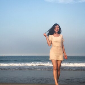 Puede incluir: Una mujer con un vestido blanco de crochet sonr&iacute;e mientras est&aacute; de pie en una playa de arena con olas del oc&eacute;ano en el fondo.