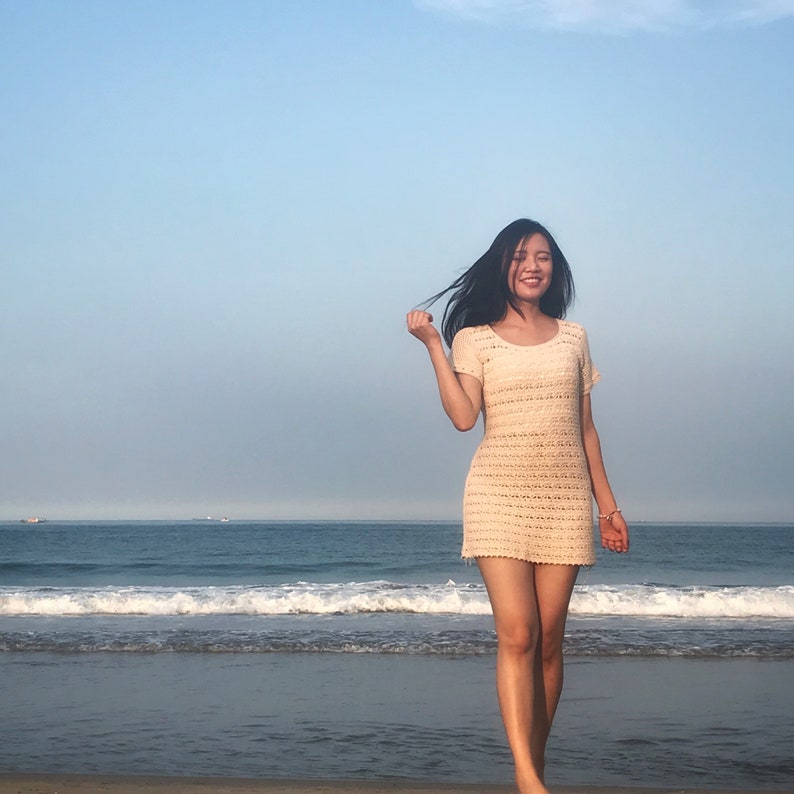 Puede incluir: Una mujer con un vestido blanco de crochet sonr&iacute;e mientras est&aacute; de pie en una playa de arena con olas del oc&eacute;ano en el fondo.