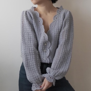 May include: A pale blue crochet cardigan with a scalloped neckline and cuffs. The long-sleeved sweater has a button-up front and a delicate, open-weave pattern. The cardigan is a soft, pastel colour.