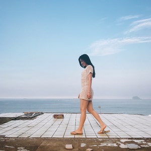 Puede incluir: Una mujer con un vestido de crochet blanco camina por un patio de baldosas con vistas al oc&eacute;ano. El cielo es azul y el agua est&aacute; tranquila.