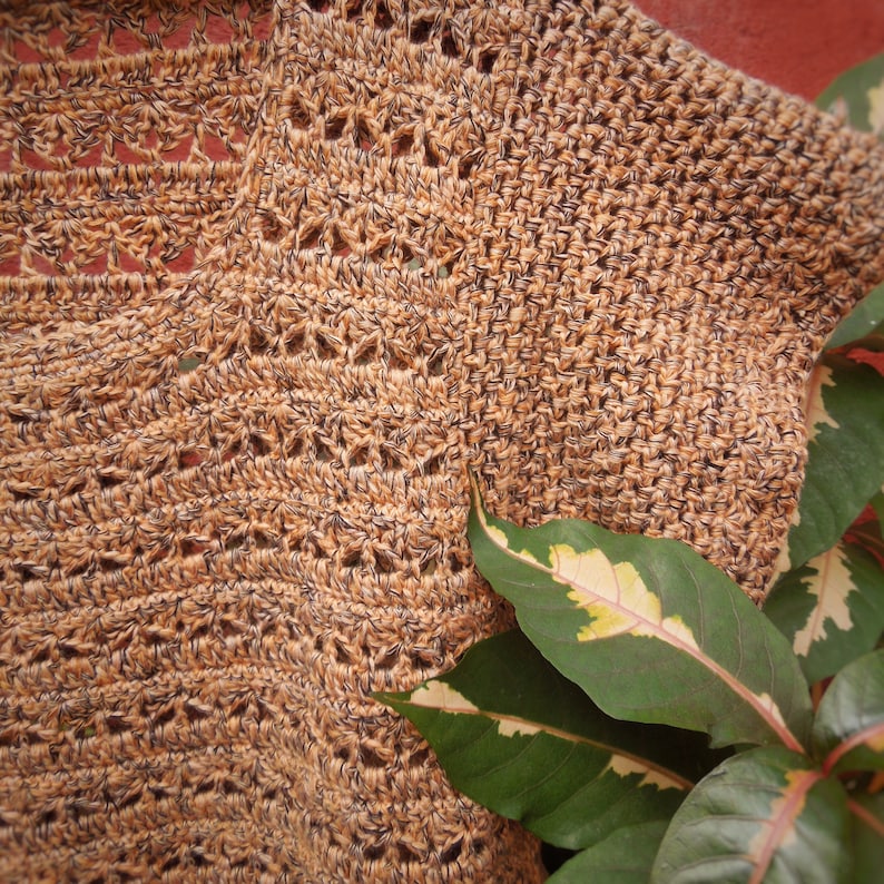 Puede incluir: Primer plano de una tela de crochet marr&oacute;n y negro con un patr&oacute;n texturizado. Una planta verde con variegaci&oacute;n blanca es parcialmente visible en la esquina inferior derecha de la imagen.