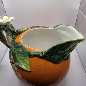 Könnte beinhalten: Ein Keramikkrug in Form einer texturierten Orange mit glänzender Oberfläche. Der Griff ist als grüner Zweig mit dunkelgrünen Blättern gestaltet, an dessen oberem Ende nahe dem Rand eine weiße Blüte mit gelber Mitte befestigt ist.