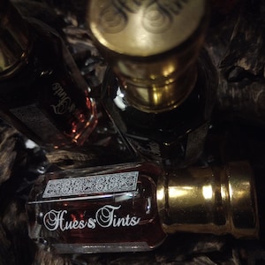 Puede incluir: Tres frascos de perfume de vidrio con tapones dorados y líquido oscuro. Las botellas llevan la inscripción "Hues & Tints" en una elegante escritura. Están rodeadas de trozos de madera oscura, creando una estética lujosa.