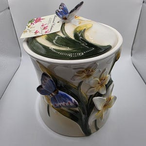 Może przedstawiać: Biały ceramiczny pojemnik z pokrywką zwieńczoną niebieskim motylem. Korpus ozdobiony jest wypukłymi, trójwymiarowymi zielonymi liśćmi, białymi i żółtymi kwiatami lilii oraz niebieskim motylem. Etykieta zawiera napis Botanical Collection.