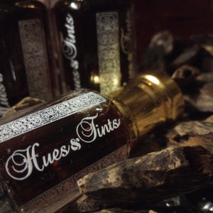 Puede incluir: Tres pequeñas botellas de vidrio con tapas doradas y un líquido marrón oscuro en su interior. Las botellas están etiquetadas con "Hues & Tints" en letras blancas. Las botellas están sobre un montón de astillas de madera marrón.
