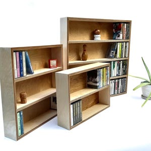 May include: Three wooden shelves with different sizes, each filled with books and DVDs. The shelves are made of light wood with a natural finish. The shelves are standing on a white surface.
