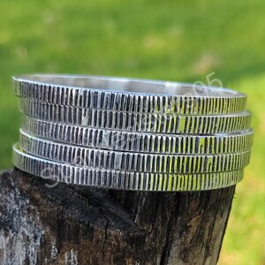 Puede incluir: Una pila de cinco brazaletes plateados con un diseño texturizado y acanalado. Los brazaletes se exhiben sobre un trozo de madera contra un fondo verde borroso. Los brazaletes parecen ser de metal.