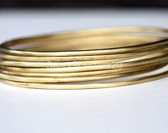 Puede incluir: Un conjunto de ocho pulseras de metal dorado apiladas unas sobre otras. Las pulseras tienen todas el mismo ancho y un acabado liso y pulido.