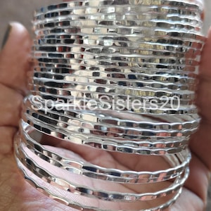 Pode incluir: Uma pilha de 12 pulseiras de prata martelada. As pulseiras são finas e têm uma superfície texturizada.