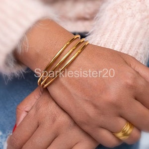 Op de afbeelding: Een set van vier gouden bangle armbanden gestapeld op de pols van een persoon. De armbanden zijn glad en glanzend.