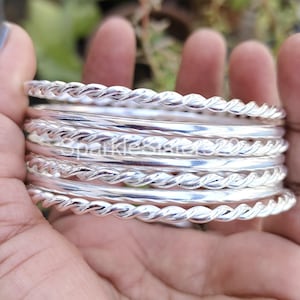 May include: A set of eight silver bangles with a twisted rope design. The bangles are stacked on top of each other and are held in a hand.