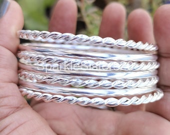 Pode incluir: Um conjunto de oito pulseiras de prata com um design de corda trançada. As pulseiras estão empilhadas umas sobre as outras e são seguradas na mão.