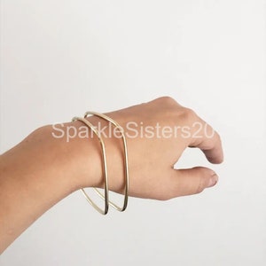 Puede incluir: Dos pulseras de metal dorado apiladas en la mu&ntilde;eca de una persona. Las pulseras son simples y de forma geom&eacute;trica.