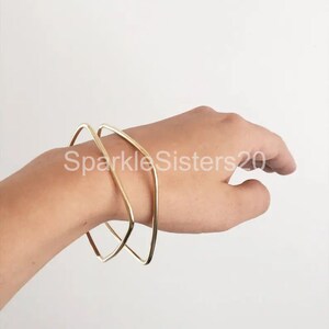 Puede incluir: Dos pulseras de brazalete geom&eacute;tricas doradas apiladas en una mu&ntilde;eca. Las pulseras est&aacute;n abiertas y tienen una forma rectangular ligeramente redondeada.