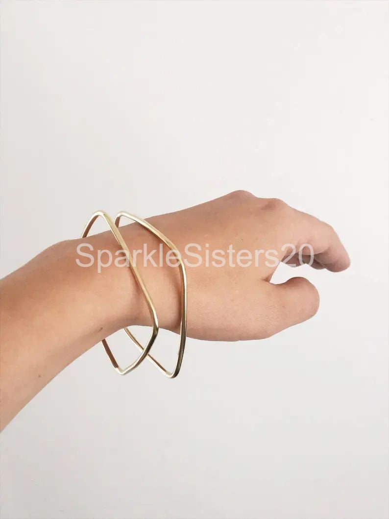 Puede incluir: Dos pulseras de brazalete geom&eacute;tricas doradas apiladas en una mu&ntilde;eca. Las pulseras est&aacute;n abiertas y tienen una forma rectangular ligeramente redondeada.