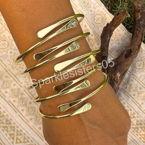 May include: A set of five gold-toned metal bangles with a unique, angular design. The bangles are stacked on a person's arm.
