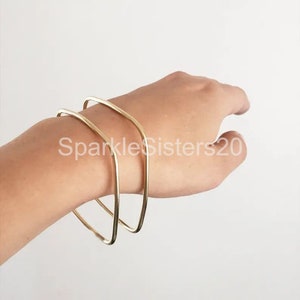Puede incluir: Dos pulseras de metal dorado apiladas en la mu&ntilde;eca de una persona. Las pulseras son simples y geom&eacute;tricas, con una forma ligeramente cuadrada.