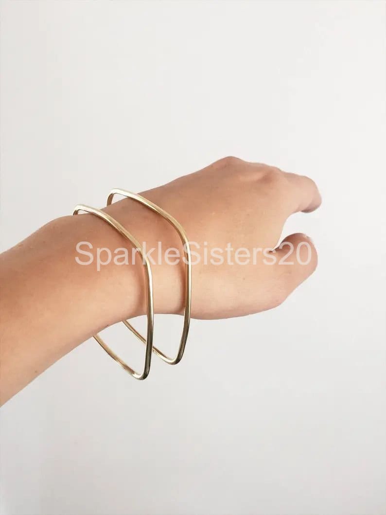 Puede incluir: Dos pulseras de metal dorado apiladas en la mu&ntilde;eca de una persona. Las pulseras son simples y geom&eacute;tricas, con una forma ligeramente cuadrada.
