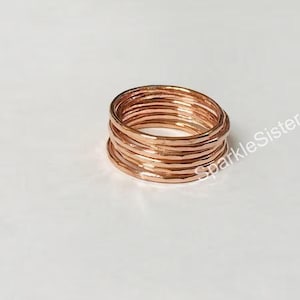 Op de afbeelding: Een stapel van zeven dunne, roségoudkleurige metalen ringen met een gehamerde, getextureerde afwerking. De ringen zijn in een cilindrische vorm gerangschikt. De tekst SparkleSisters05 is zichtbaar in witte letters aan de rechterkant.