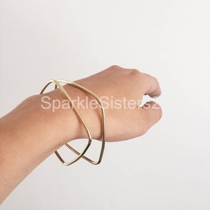 Puede incluir: Dos pulseras geom&eacute;tricas doradas apiladas en una mu&ntilde;eca. Las pulseras est&aacute;n hechas de alambre grueso y redondeado y tienen un dise&ntilde;o simple y moderno.
