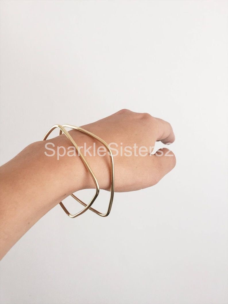Puede incluir: Dos pulseras geom&eacute;tricas doradas apiladas en una mu&ntilde;eca. Las pulseras est&aacute;n hechas de alambre grueso y redondeado y tienen un dise&ntilde;o simple y moderno.
