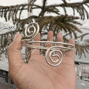 May include: Silver-toned arm cuff with spiral designs at each end. The cuff is held up against a backdrop of green foliage. The design features two large, open spirals connected by a smooth, curved band.