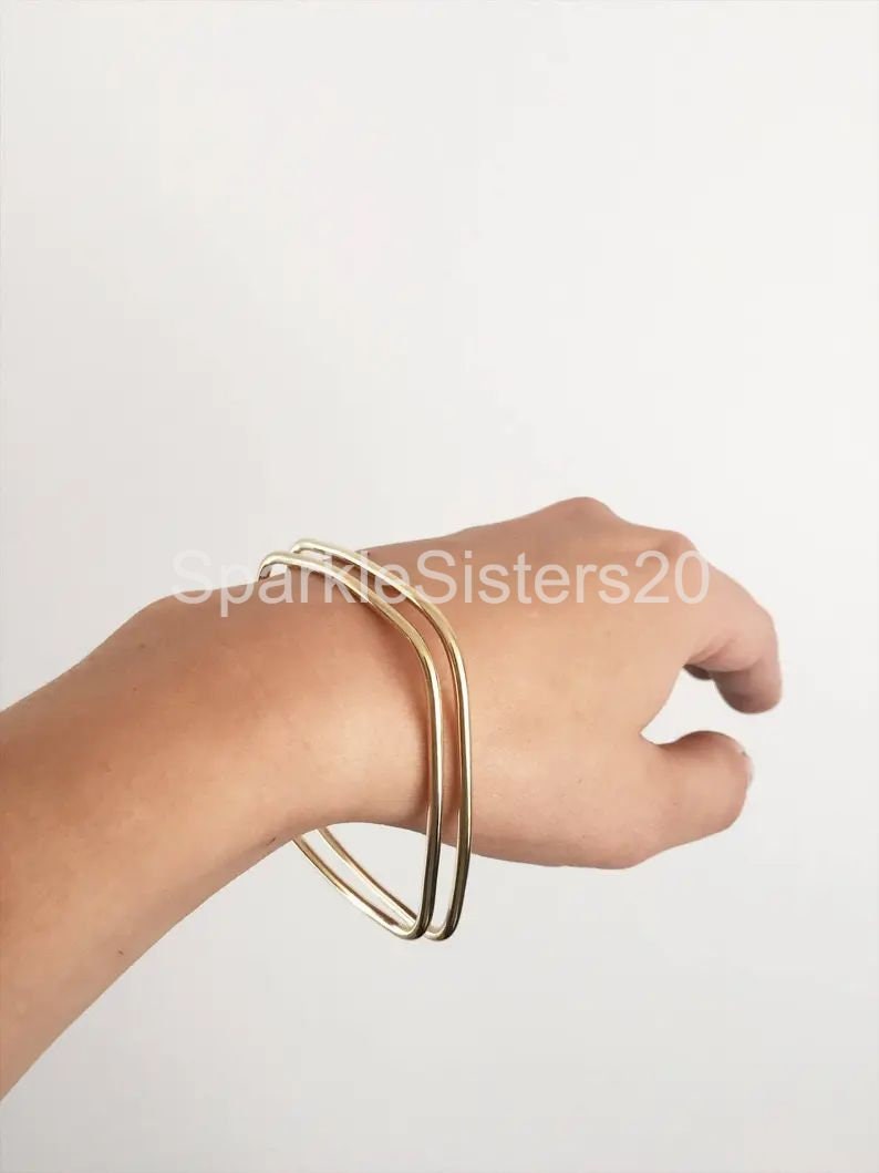 Puede incluir: Dos pulseras doradas apiladas en la mu&ntilde;eca de una persona. Las pulseras son simples y tienen forma cuadrada.